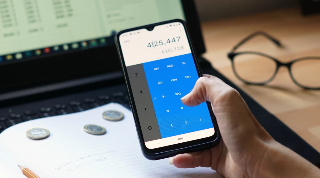 Person using a calculator app on a smartphone while working at a desk with a laptop, notebook, coins, and glasses nearby.