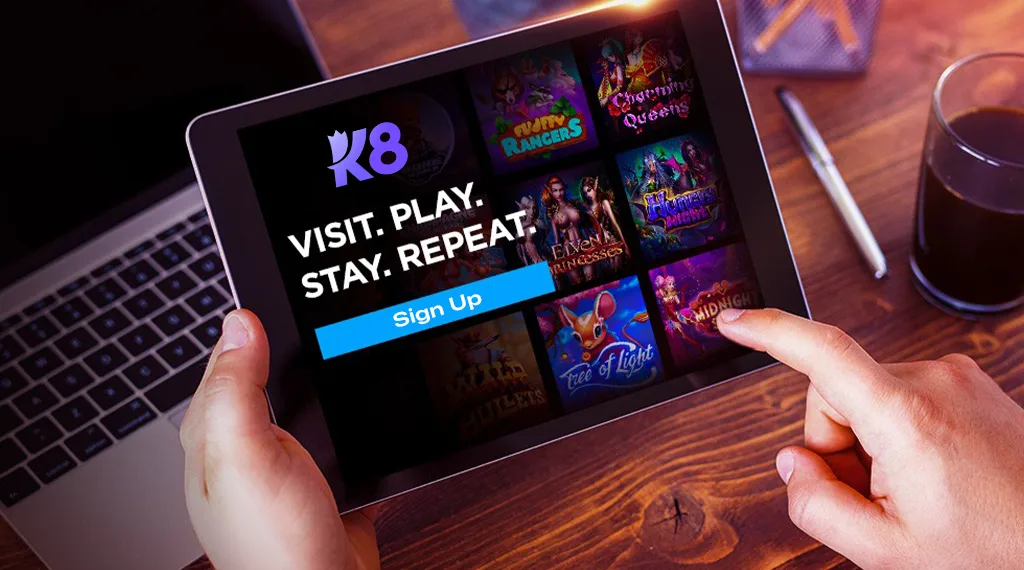 A person using a tablet to sign up for K8 online gaming, showing a "Visit. Play. Stay. Repeat." message with a selection of game thumbnails.