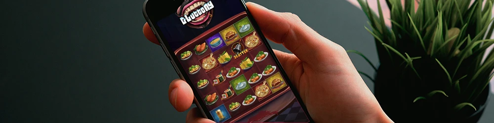 A person holding a smartphone displaying the Gluttony slot game with various food symbols like burgers, pizza, and drinks on the reels.