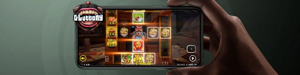 A person holding a smartphone showing the Gluttony slot game with food-themed symbols like burgers, drinks, and multiplier features displayed on the reels.